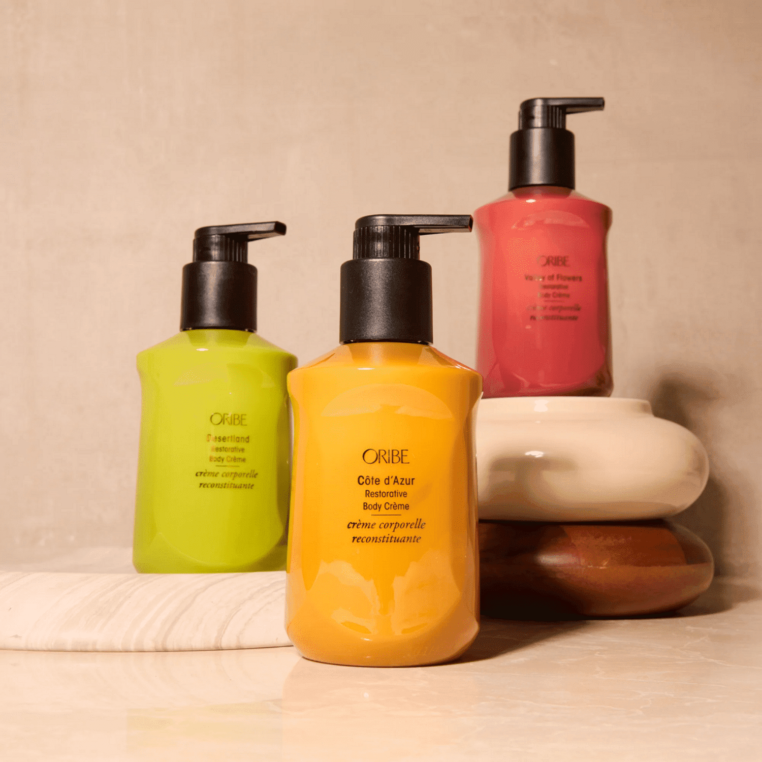 Three Oribe body creams in green, yellow, and pink bottles on elegant soapstone display.