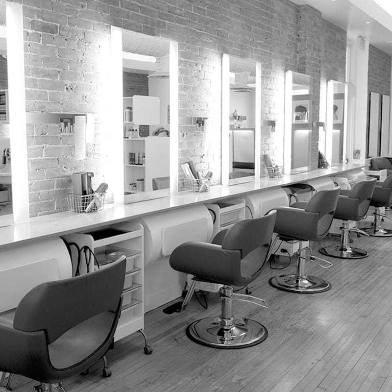Modern hair salon with lit mirrors, empty chairs, and brick wall.